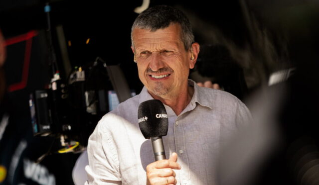 Male presenter holding a Canal+ microphone in front of camera gear during a live interview, wearing a light button-up shirt and squinting slightly in bright light.