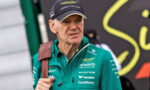 Senior man wearing an Aston Martin Formula One Team cap and green sponsor jacket, looking to the side, with a brown shoulder strap visible across his chest.