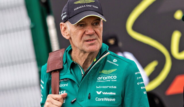 Senior man wearing an Aston Martin Formula One Team cap and green sponsor jacket, looking to the side, with a brown shoulder strap visible across his chest.