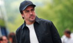 Man in a black cap and jacket, white tee, looking to the left outdoors.