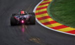Formula race car navigating a sharp turn on a dark asphalt track with red and yellow striped runoff curbs on the inside edge and green grass beyond.