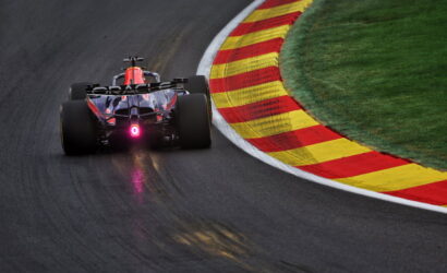 Formula race car navigating a sharp turn on a dark asphalt track with red and yellow striped runoff curbs on the inside edge and green grass beyond.