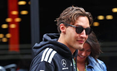Young man in a dark hoodie with Mercedes‑AMG logos and sunglasses, standing outdoors with a woman in the background.