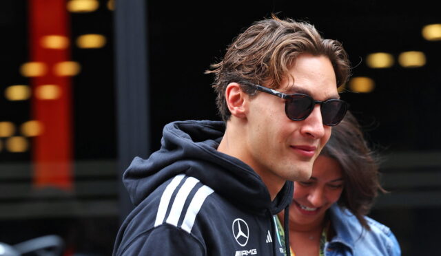 Young man in a dark hoodie with Mercedes‑AMG logos and sunglasses, standing outdoors with a woman in the background.