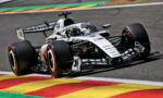 White Formula 1 race car navigating a turn on a track, driver in helmet visible, beside colorful painted curbs.