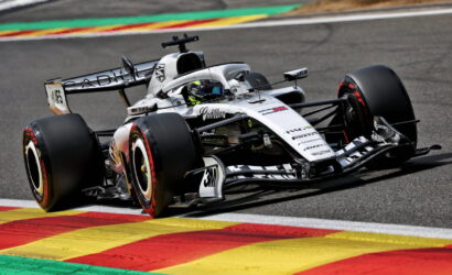White Formula 1 race car navigating a turn on a track, driver in helmet visible, beside colorful painted curbs.