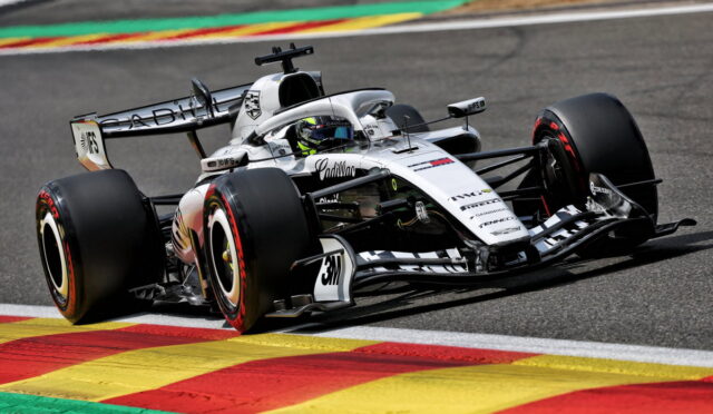 White Formula 1 race car navigating a turn on a track, driver in helmet visible, beside colorful painted curbs.