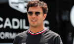 Man wearing sunglasses and a black T-shirt with Cadillac logos, standing outdoors at a car event with a blurred Cadillac sign in the background