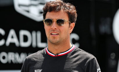 Man wearing sunglasses and a black T-shirt with Cadillac logos, standing outdoors at a car event with a blurred Cadillac sign in the background
