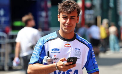 Young man in a blue-and-white racing jersey outdoors, looking at his smartphone while holding a water bottle.