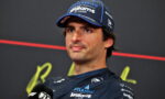 F1 driver in a Williams cap speaks at a press conference, microphone in hand and dark backdrop behind him.