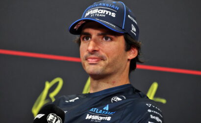 F1 driver in a Williams cap speaks at a press conference, microphone in hand and dark backdrop behind him.
