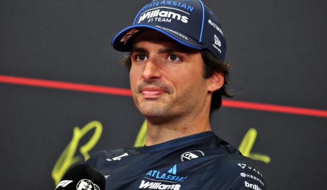 F1 driver in a Williams cap speaks at a press conference, microphone in hand and dark backdrop behind him.