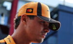 Close-up of a man wearing an orange racing cap with Mastercard logo and Monster energy visor, looking to the side in a pit area