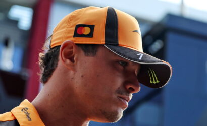 Close-up of a man wearing an orange racing cap with Mastercard logo and Monster energy visor, looking to the side in a pit area