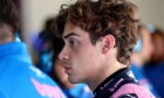 Side profile of a young man with tousled brown hair wearing a pink racing suit in a garage setting.
