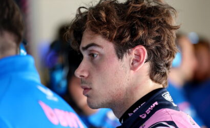 Side profile of a young man with tousled brown hair wearing a pink racing suit in a garage setting.