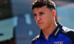 Young man in a blue racing polo with sponsor logos, looking serious outdoors with blurred background.