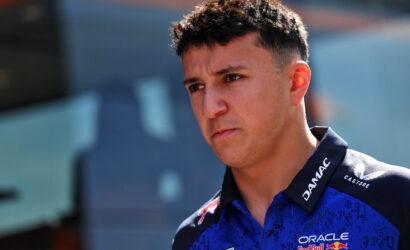 Young man with short dark hair in a blue racing polo with sponsor logos, looking pensively to the left outdoors.