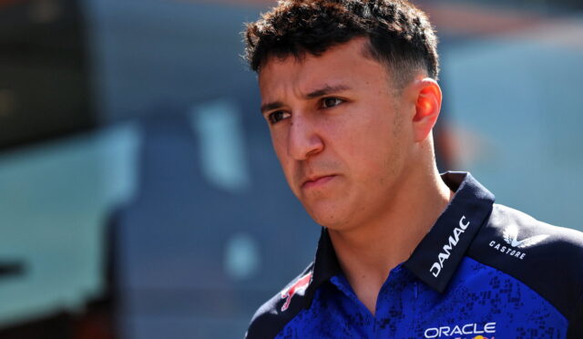 Young man with short dark hair in a blue racing polo with sponsor logos, looking pensively to the left outdoors.