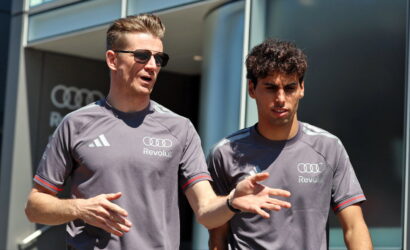 Two men in gray Revolut shirts stand outside a modern glass building; one wearing sunglasses speaks and gestures while the other listens.