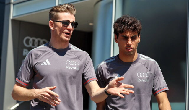 Two men in gray Revolut shirts stand outside a modern glass building; one wearing sunglasses speaks and gestures while the other listens.