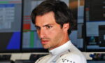 Formula 1 driver in a white racing suit looks focused in a control room with multiple monitors in the background.