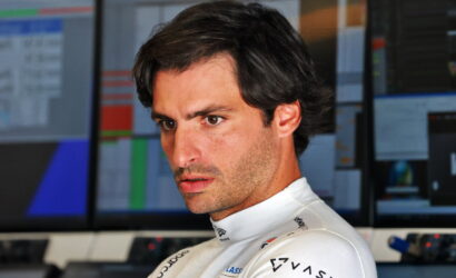 Formula 1 driver in a white racing suit looks focused in a control room with multiple monitors in the background.