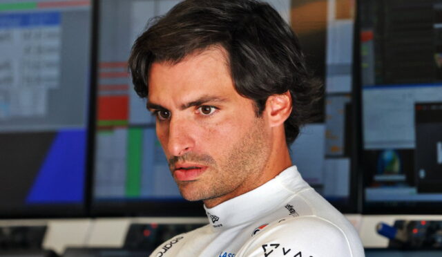 Formula 1 driver in a white racing suit looks focused in a control room with multiple monitors in the background.
