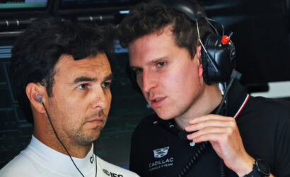 Two men with headsets in a racing garage, one in a white team shirt and the other in a black Cadill lac polo, consulting intently.