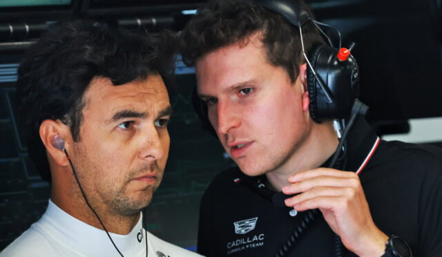 Two men with headsets in a racing garage, one in a white team shirt and the other in a black Cadill lac polo, consulting intently.
