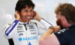 Race car driver in a white Barclays racing suit smiling as a team member adjusts his ear area before a race.