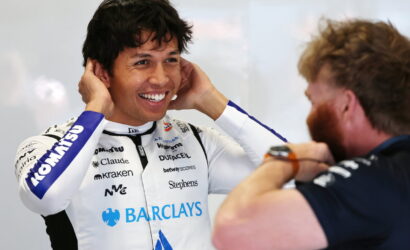 Race car driver in a white Barclays racing suit smiling as a team member adjusts his ear area before a race.