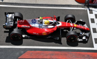 Red Ferrari Formula 1 car (#44) on a pit lane/track surface with a yellow-helmeted driver inside the cockpit, sponsor logos visible along the body.