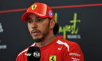 Male Formula 1 driver in a red Ferrari cap and team shirt giving an interview, holding a FIA branded microphone against a dark backdrop with sponsor logos.