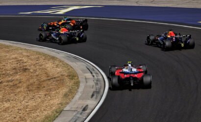 Multiple Formula 1 cars race around a sharp curve on a dark asphalt track, with a red and blue car leading from the foreground and several others close behind.