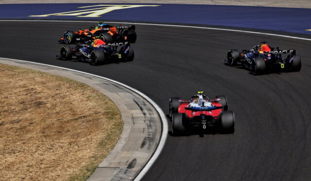 Multiple Formula 1 cars race around a sharp curve on a dark asphalt track, with a red and blue car leading from the foreground and several others close behind.