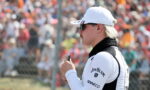 Side profile of a man in a white cap and sunglasses, holding a small device at a crowded motorsport event behind a fence.