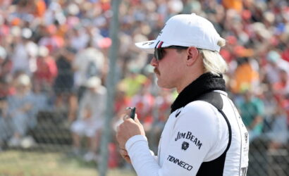 Side profile of a man in a white cap and sunglasses, holding a small device at a crowded motorsport event behind a fence.