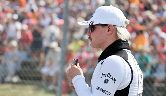 Side profile of a man in a white cap and sunglasses, holding a small device at a crowded motorsport event behind a fence.