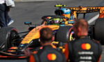 Orange Formula 1 car in the pit lane with a helmeted driver in the cockpit and blurred crew in the foreground.