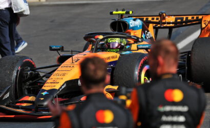 Orange Formula 1 car in the pit lane with a helmeted driver in the cockpit and blurred crew in the foreground.