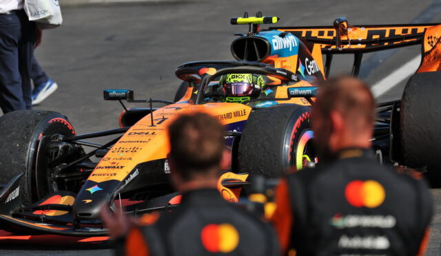 Orange Formula 1 car in the pit lane with a helmeted driver in the cockpit and blurred crew in the foreground.