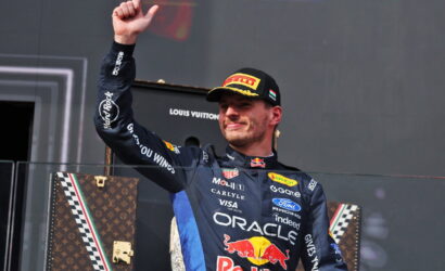 Racing driver on a podium raises a fist, wearing a dark racing suit with Red Bull and Oracle logos and a cap.