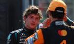 Two race car drivers in team suits chat in a pit area, one facing the other while the other adjusts his helmet strap.