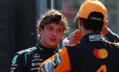 Two race car drivers in team suits chat in a pit area, one facing the other while the other adjusts his helmet strap.