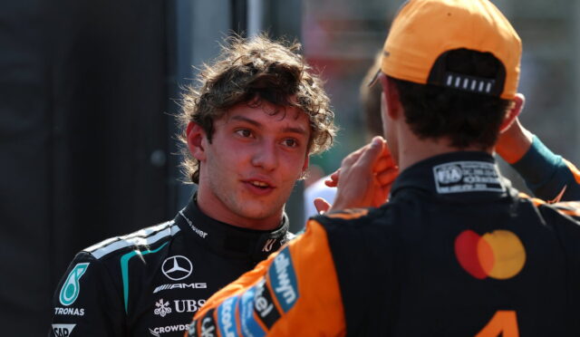 Two race car drivers in team suits chat in a pit area, one facing the other while the other adjusts his helmet strap.