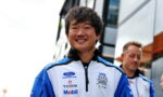 Smiling young man in a blue-and-white racing jacket with sponsor logos stands outdoors, another person in background.
