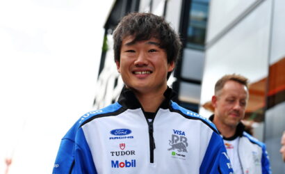 Smiling young man in a blue-and-white racing jacket with sponsor logos stands outdoors, another person in background.