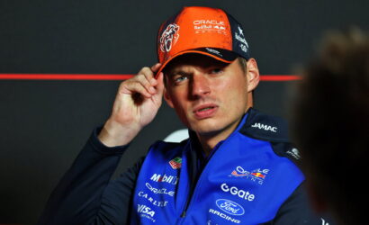 Racing driver in a blue Oracle Red Bull Racing jacket adjusts an orange cap, facing camera against a dark background.